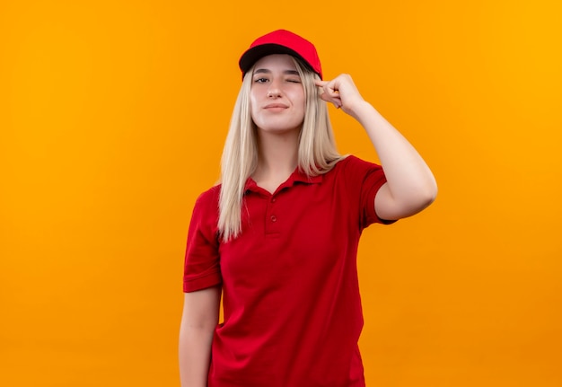 빨간 t- 셔츠와 모자를 쓰고 깜박이 배달 젊은 여자는 고립 된 주황색 벽에 머리에 그녀의 손가락을 넣어