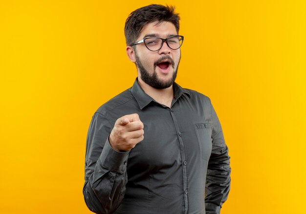 Blinked joyful young businessman wearing glasses showing you gesture isolated on yellow background