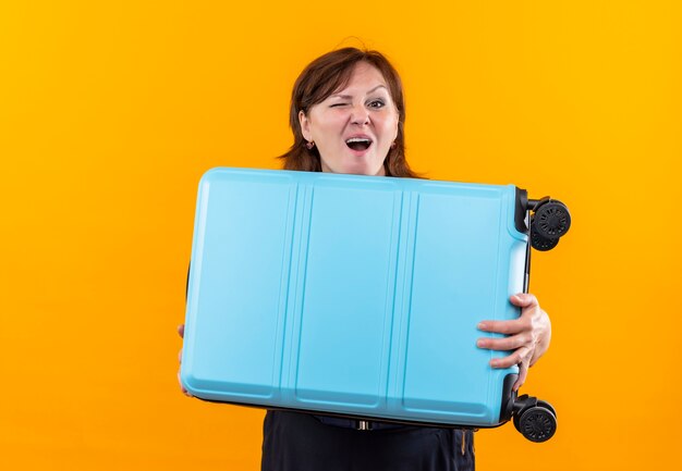 Blinked middle-aged traveler woman holding suitcase on isolated yellow wall