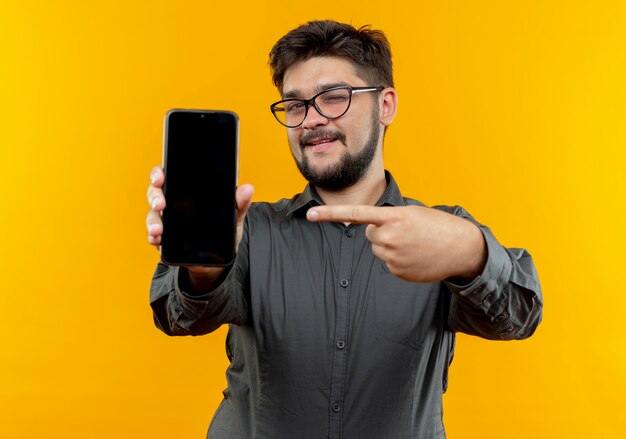 Blinked pleased young businessman wearing glasses holding and points at phone isolated on yellow background