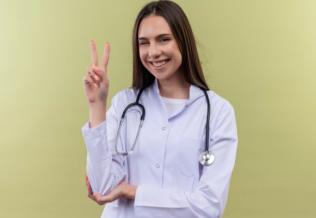 Blinked smiling young doctor girl wearing stethoscope medical gown showing peace gesture on green wall