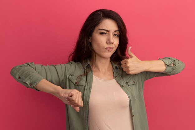 Blinked young beautiful girl wearing olive green t-shirt showing thumbs up and down isolated on pink wall