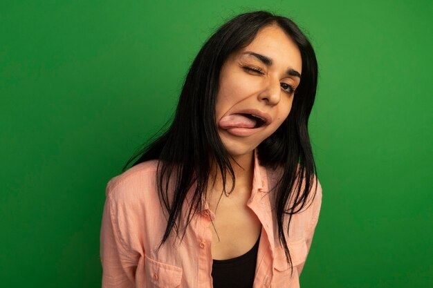 Blinked young beautiful girl wearing pink t-shirt showing tongue isolated on green