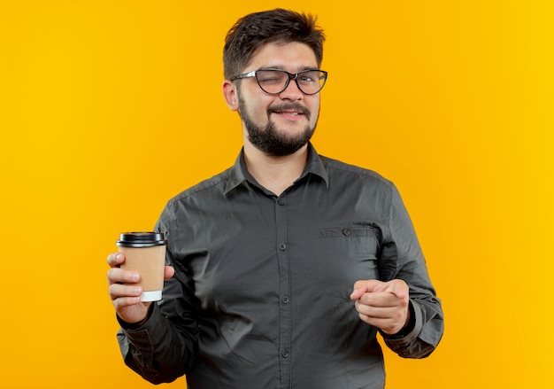 Blinked young businessman wearing glasses holding cup of coffee and showing you gesture isolated on yellow background
