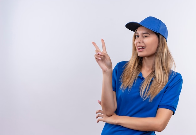 Blinked young delivery girl wearing blue uniform and cap showing peace gesture isolated on white wall with copy space