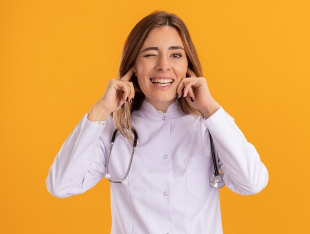 Blinked young female doctor wearing medical robe with stethoscope closed ears isolated on yellow wall