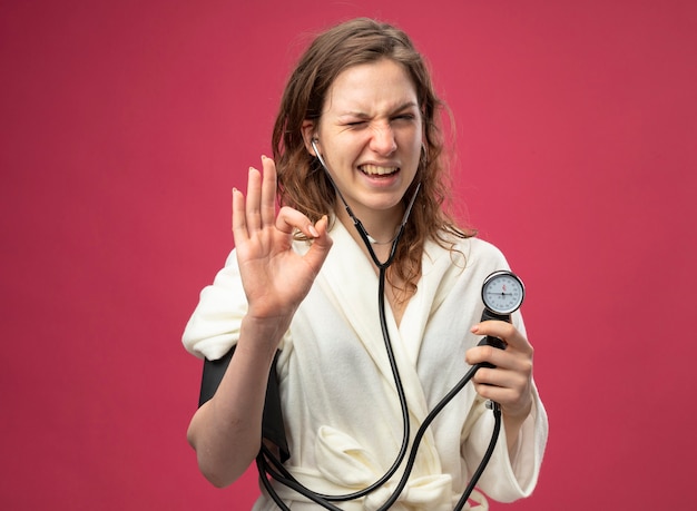 Blinked young ill girl wearing white robe measuring her own pressure with sphygmomanometer showing okay gesture isolated on pink