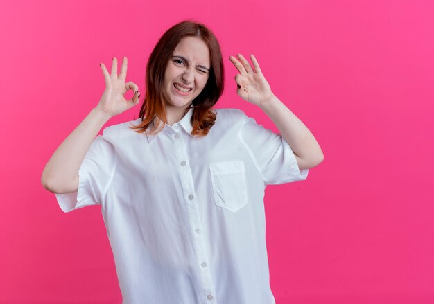 Blinked young redhead girl showing okey gesture isolated on pink wall with copy space