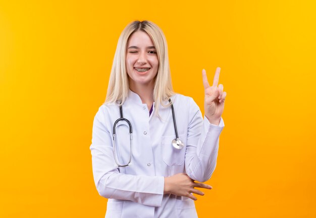 Мигающая молодая девушка-врач со стетоскопом в медицинском халате и стоматологической скобе, показывающая жест мира на изолированной желтой стене