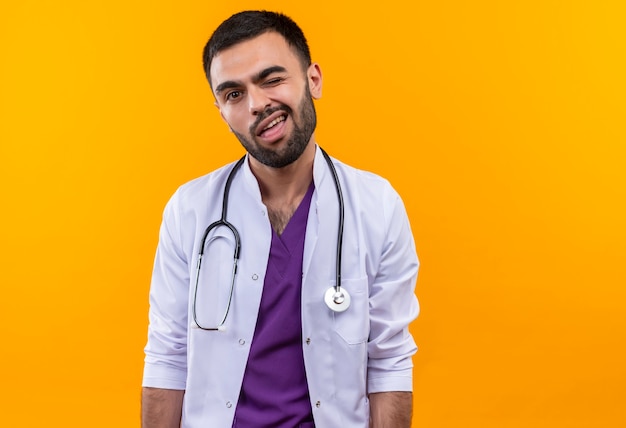 Blinking young male doctor wearing stethoscope medical gown on isolated yellow wall