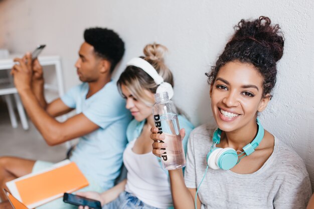 Blissful african girl holding bottle of water and posing with smile with friends. Portrait of curly female student , blonde young woman and black guy making photo.