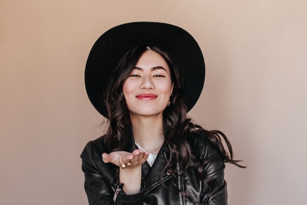 베이지 색 배경에 몸짓 모자에 행복 한 아시아 여자. 가죽 재킷에 로맨틱 곱슬 일본 여자의 스튜디오 샷.