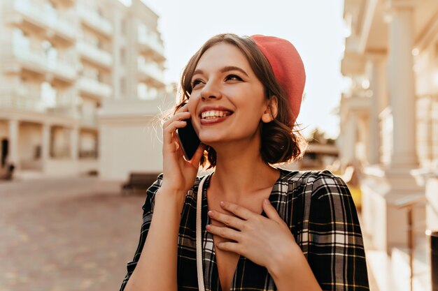 電話で話しているベレー帽の至福の美しい少女。スマートフォンを持って通りを歩いている魅力的な短髪の女性。