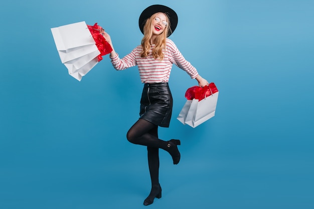 Free photo blissful blonde girl in leather skirt dancing on blue wall.  joyful young woman with shopping bags.