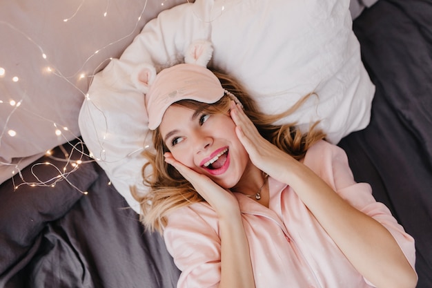 驚きの笑顔でベッドでポーズをとる至福のブロンドの女の子。ポジティブな感情を表現するパジャマとアイマスクで幸せな女性の屋内の肖像画。