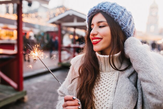 Beata donna dai capelli castani con un sorriso sincero godendo le vacanze di natale e in posa con sparkler. affascinante ragazza in morbido cappello blu che tiene la luce del bengala sulla strada.