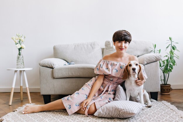 Blissful brunette girl in cute outfit sits on carpet in front of gray sofa with her puppy