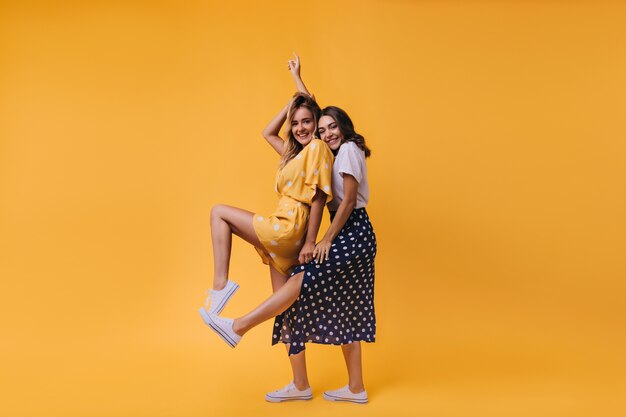 Blissful brunette girl in long skirt posing with her sister. Indoor portrait of spectacular female friends isolated on yellow.