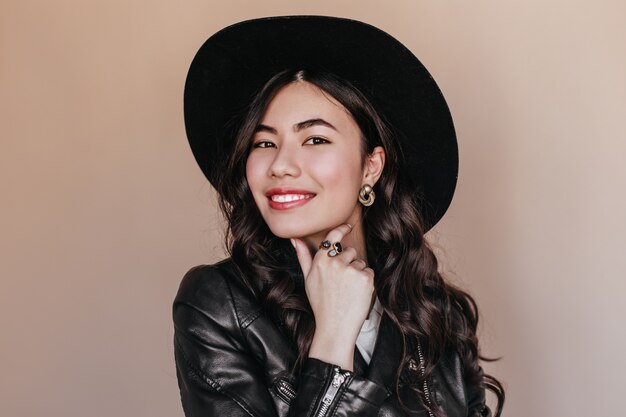Blissful chinese young woman smiling at camera. Studio shot of curly japanese woman in hat and leather jacket.