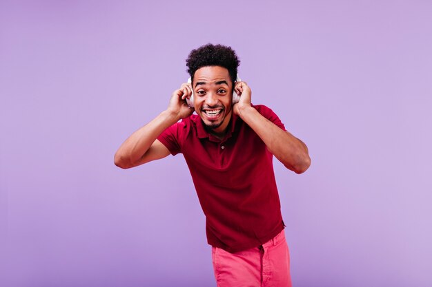 Blissful curly funny man looking. African guy in red clothes posing in white headphones.