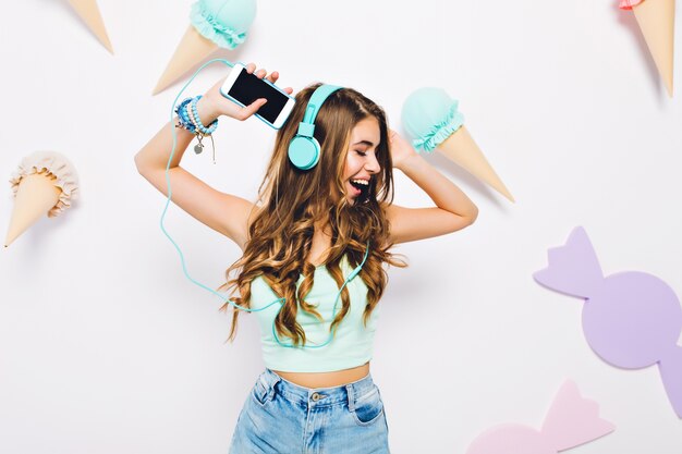 보라색 사탕과 아이스크림으로 장식 된 벽에 춤을 추는 큰 파란색 헤드폰에 행복한 곱슬 소녀. 재미와 눈으로 음악을 즐기는 쾌활 한 젊은 여자의 초상화를 폐쇄.