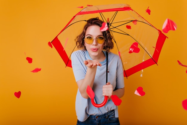 Blissful curly girl in glasses sending air kiss, standing under umbrella. Happy brunette young woman posing on yellow wall with red hearts behind.