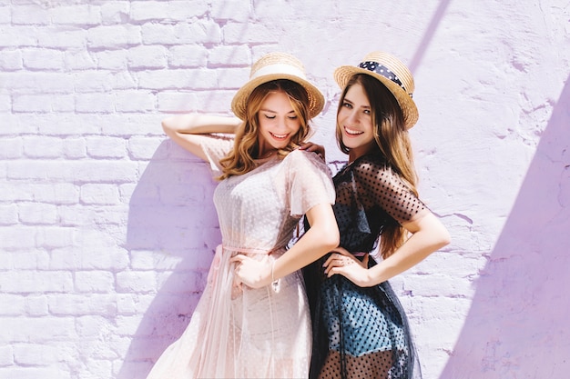 Free photo blissful curly girl in light vintage dress looking down while posing next to adorable sister