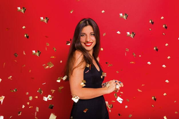 Blissful elegant stylish woman with red lipstick wearing black dress posing on red with confetti and sparks