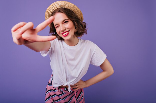 Blissful european girl with dark eyes posing in trendy straw hat. Indoor photo of sensual female model with short hair standing on purple wall and laughing.