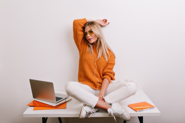 Блаженная модная женщина в модных белых штанах дурачится в офисе, сидя со скрещенными ногами за столом возле компьютера