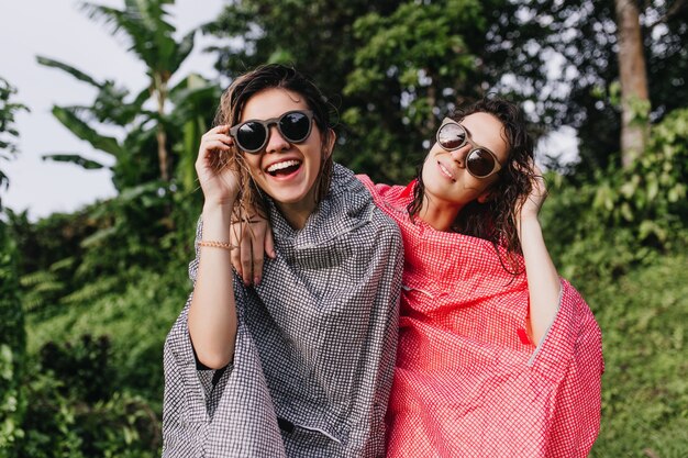 Blissful female models expressing positive emotions outdoor. Attractive women in raincoat embracing on exotic jungle.