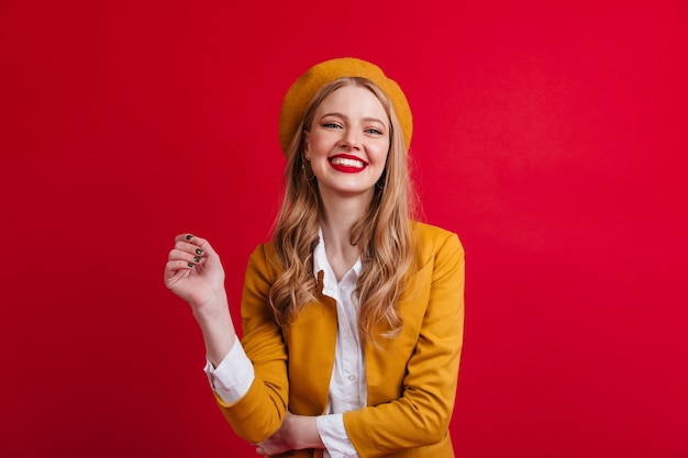 Blissful french female model laughing. Front view of blonde girl in beret isolated on red wall.