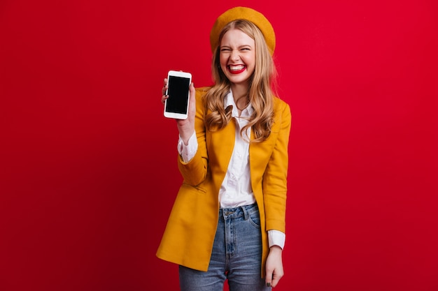 Blissful french woman holding smartphone with blank screen. Front view of elegant blonde girl in beret isolated on red wall.