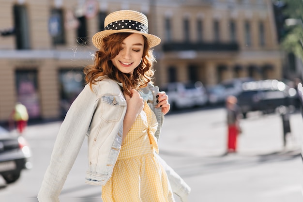 Ragazza beata dello zenzero in vestito giallo che cammina intorno alla città. ritratto all'aperto della signora caucasica felice in cappello di paglia che sorride sulla strada.