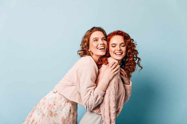 Blissful girl embracing ginger friend. Studio shot of two charming ladies posing on blue background.