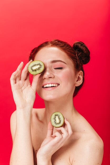 Blissful girl holding cut kiwi. Studio shot of charming ginger woman isolated on red background with fruits.