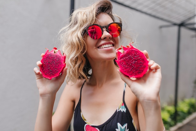 Blissful girl in sparkle sunglasses posing with red pitaya. Photo of happy european lady enjoying exotic fruits at resort.