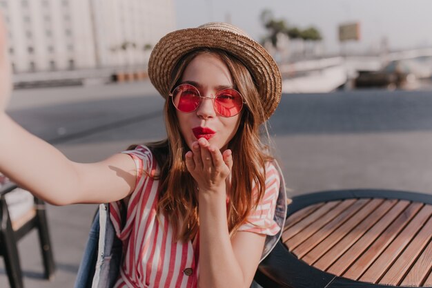 여름 날에 공기 키스를 보내는 트렌디 한 둥근 안경에 행복한 소녀. 도시에 셀카를 만드는 밀짚 모자에 매혹적인 여자의 야외 샷.