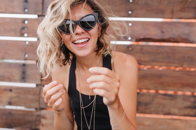 Blissful girl wears bracelet and dark sunglasses smiling during photoshoot. Portrait of gorgeous blonde female model in black attire expressing positive emotions.