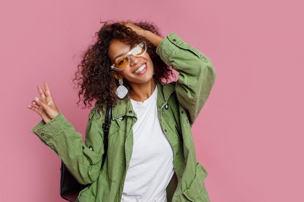 Blissful girl with African hairstyle laughing during studio photo shoot. Wearing stylish  earrings, sunglasses and green gasket. Pink background.