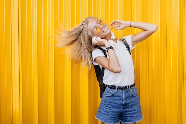 黄色の背景で踊るバックパックを持つ至福の少女ポジティブな感情を表現するデニムのショートパンツで壮大な女性モデルの屋外の肖像画