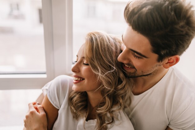 Blissful guy embracing girlfriend in morning. Indoor portrait of relaxed blonde woman chilling with boyfriend.