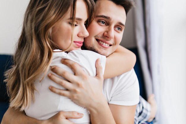 Blissful handsome man huggs his girlfriend. Indoor portrait of european married couple enjoying weekend morning.