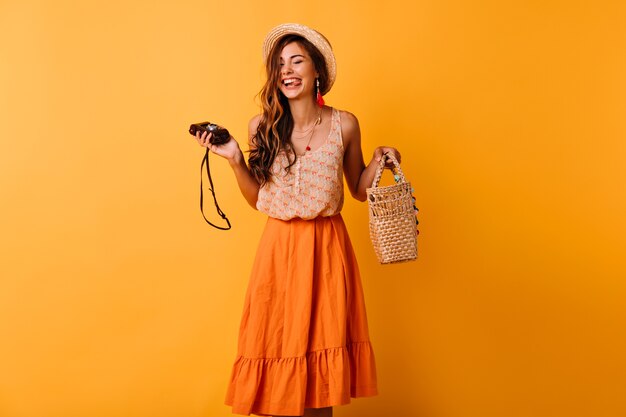 Blissful lady in trendy summer clothes posing with camera on yellow. Positive beautiful girl in hat chilling in studio.