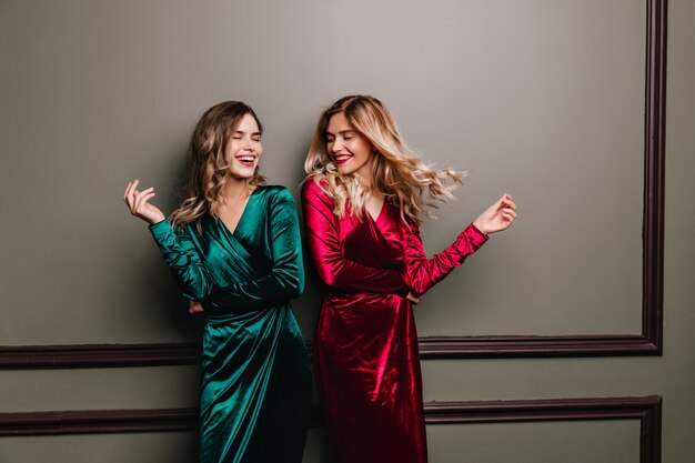 Blissful laughing girl in green dress chilling with friend. Indoor photo of two relaxed women in velvet clothes.