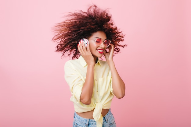 Blissful mulatto woman in yellow cotton shirt fooling around in pink room. pleased black girl with curly brown hairstyle touching white headphones and laughing.