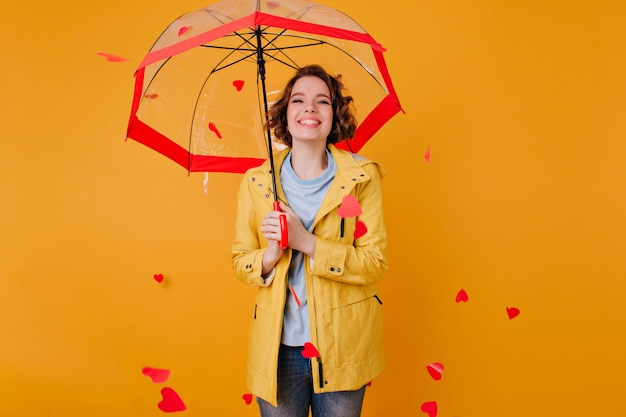 Blissful pale girl in stylish jacket standing under parasol with sincere smile. Young woman in elegant autumn attire posing on yellow wall.