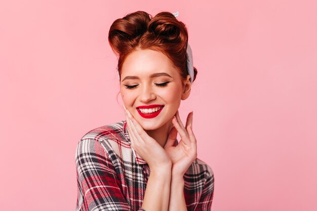 Blissful pinup woman with ginger hair smiling with closed eyes. Studio shot of refined stylish lady isolated on pink space.