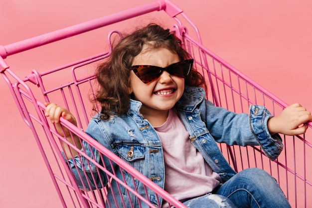 쇼핑 카트에 앉아 행복 한 유치원 아이입니다. 분홍색 배경에 재미 청바지에 웃는 아이.