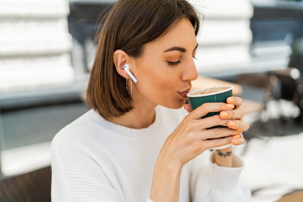 Blissful short haired woman enjoing cappucino in cafe, wearing cozy white sweater and listening to favorite music by earphones